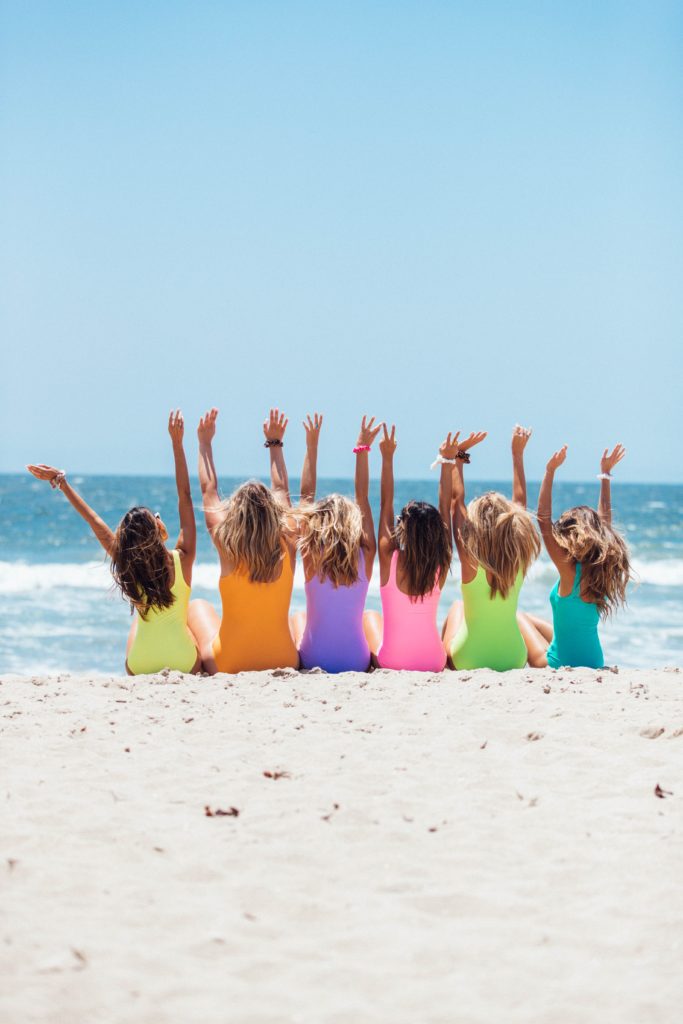 Six young women wearing different colored bathing suits sitting on the beach, celebrating with their hands in the air