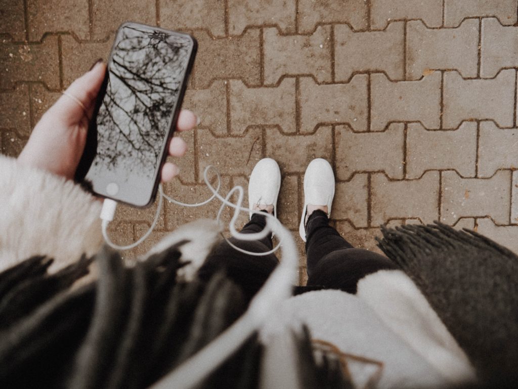 A lady with white sneakers holding a cellphone with a pair of headphones plugged in.