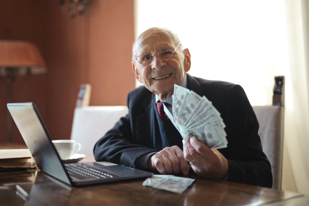On old man sitting in front of his laptop with dollar bills in his hand.
