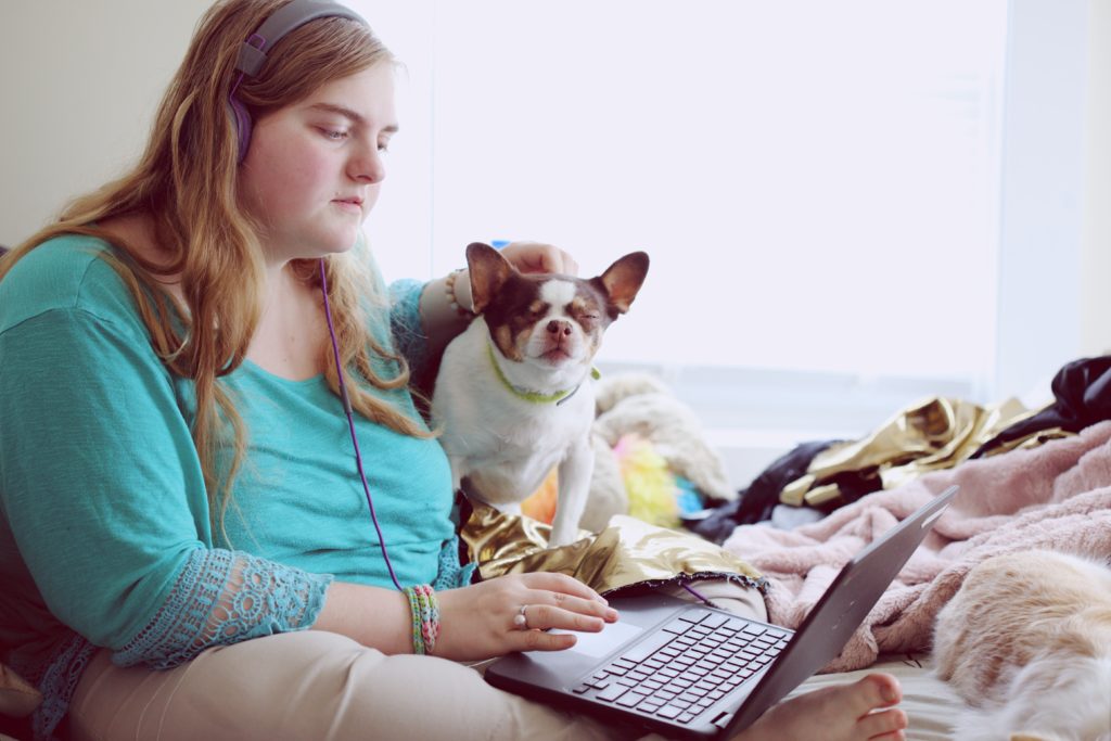 A young person in her sleepwear, on her laptop, and her dog sitting on a bed