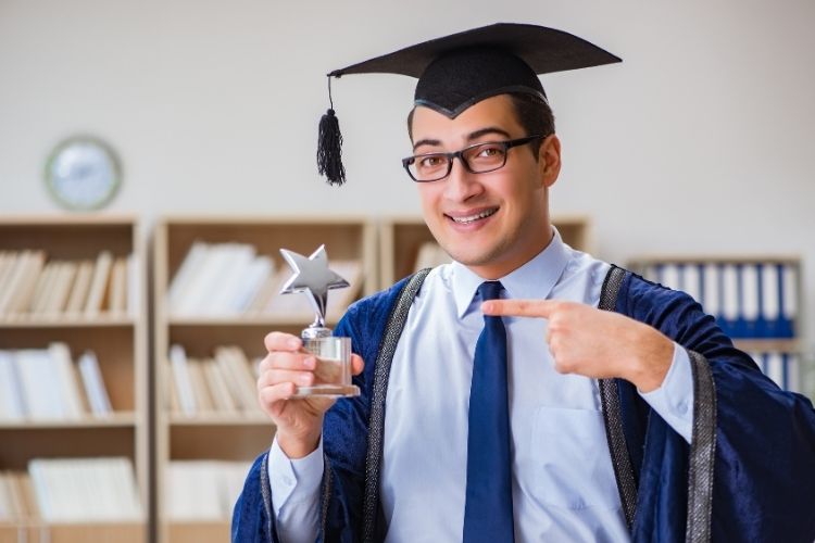 A student in a graduation gown and cap holding a trophy with a star on top.