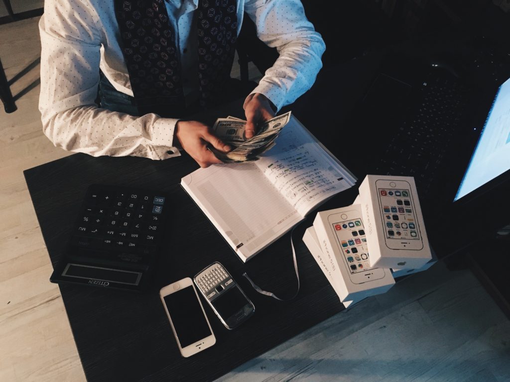 A man in a suit sitting at a desk with an open book, and a  cellphone. The man is counting money.