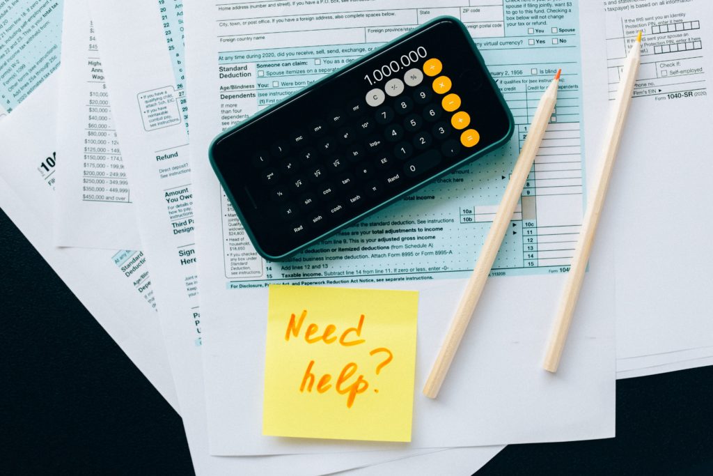 A calculator on top of a stack of documents with a sticky note saying "Need help?".