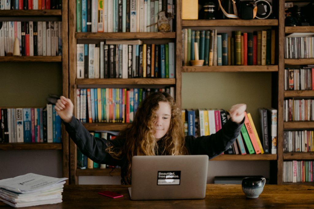 A girl in a library sitting in front of a laptop with a her arms up in the air in a victorious stance.