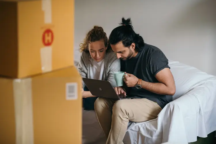 A couple sitting together, looking at a laptop with moving boxes around them.