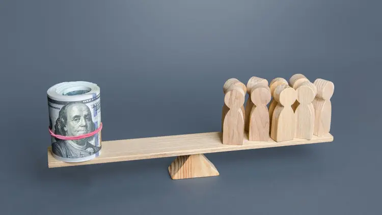 A see saw with a roll of money notes on one side, and wooden people on the other side.