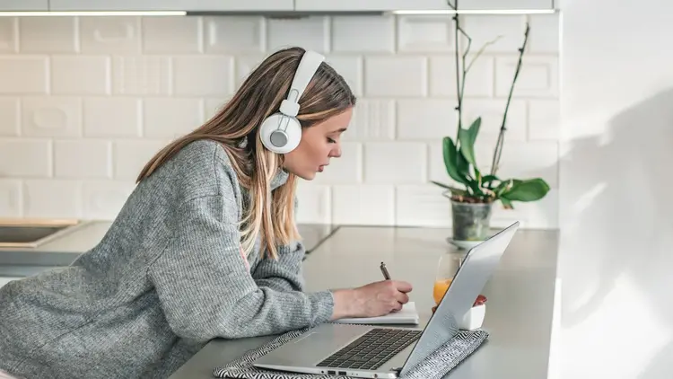 A lady standing in her kitchen with headphones, taking notes while looking at her laptop. 